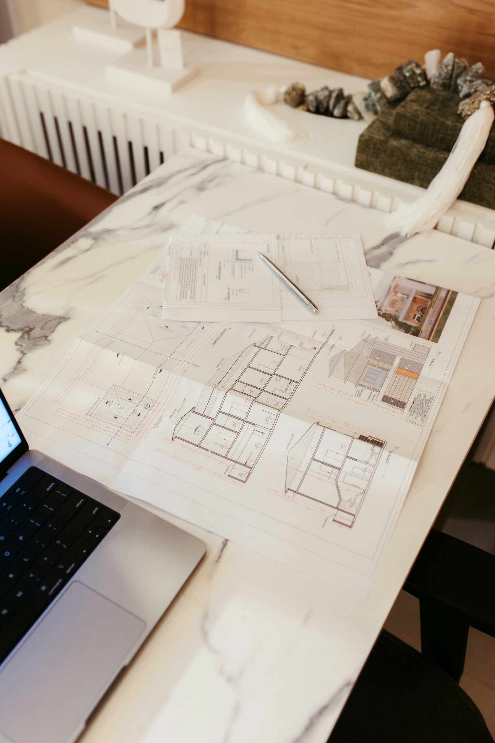 Architectural blueprints, sections, and details spread across a desk beside a laptop